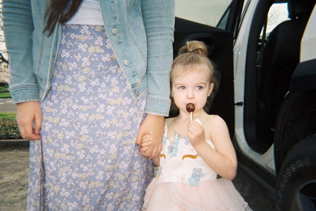 A girl with a lolipop holding the hand of an adult. Captured with a disposable camera on film.