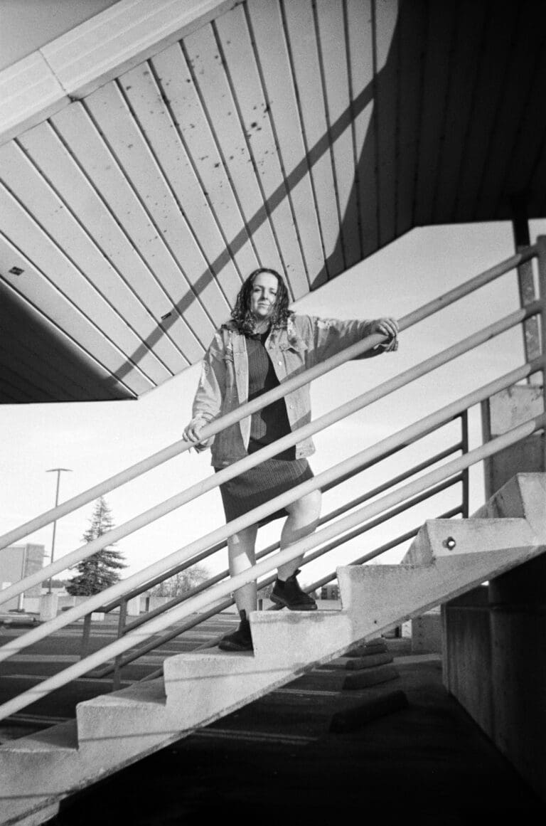 A black and white image of a girl standing on stairs, holding a railing while looking directly at the camera. Captured with film through a disposable, single-use camera.